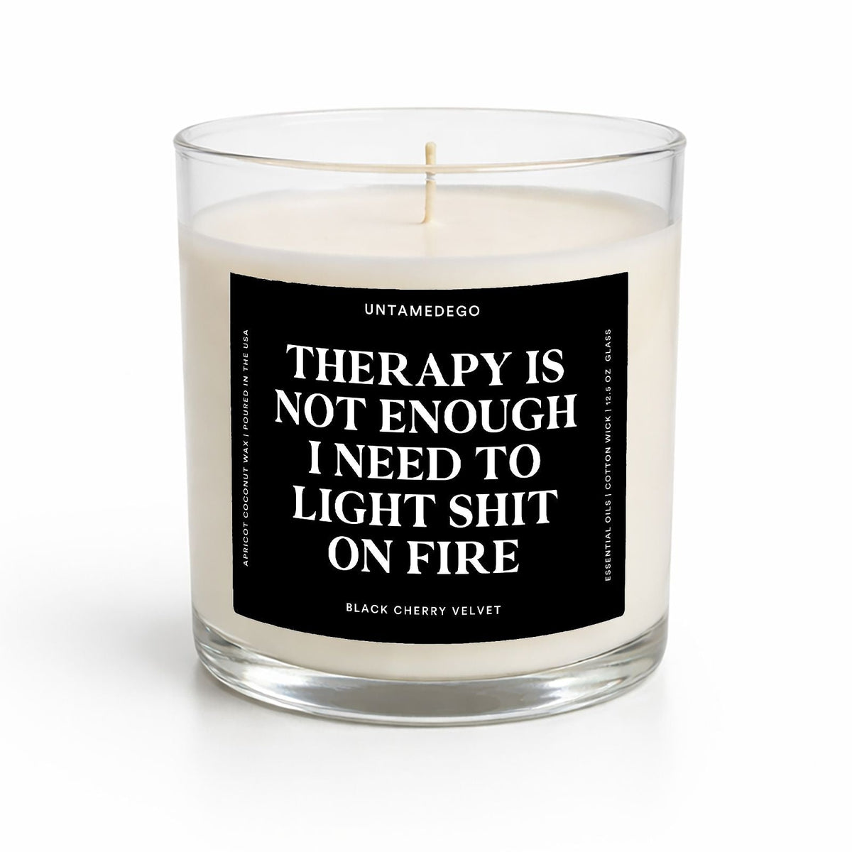 A clear glass jar candle with white wax and a black label featuring bold white text that reads, 'THERAPY IS NOT ENOUGH I NEED TO LIGHT SHIT ON FIRE.' The brand name 'UNTAMEDEGO' is visible at the top of the label, and 'ESPRESSO' appears at the bottom.