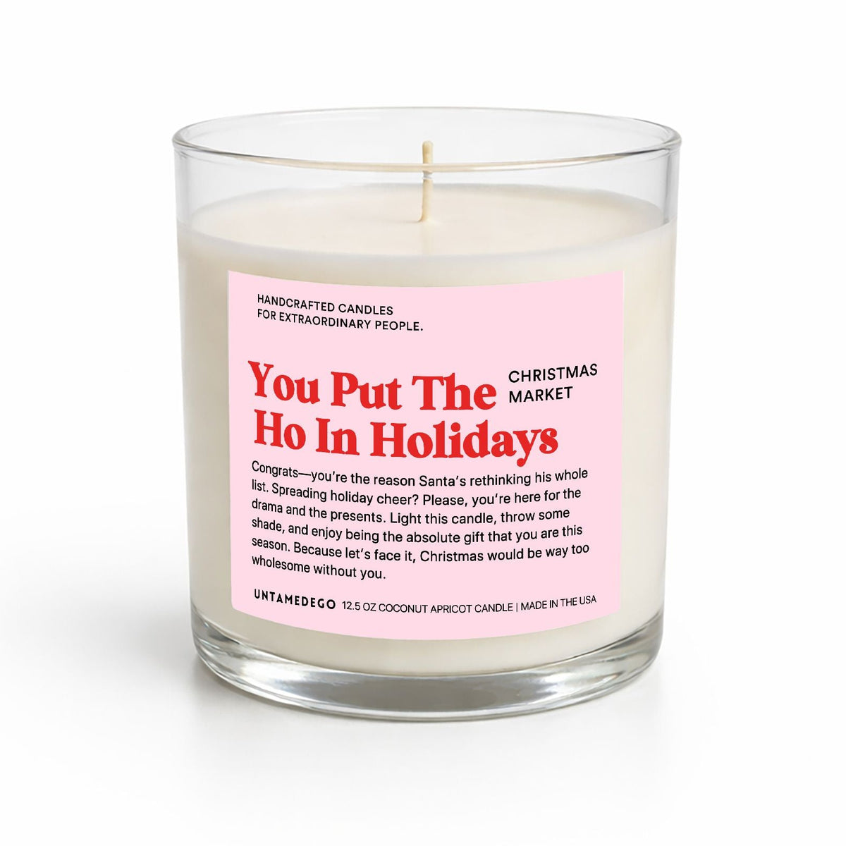 A handcrafted coconut soy wax candle in a clear glass jar with a pink and red label that reads 'You Put The Ho In Holidays' by Unnamed Co., on a white background.