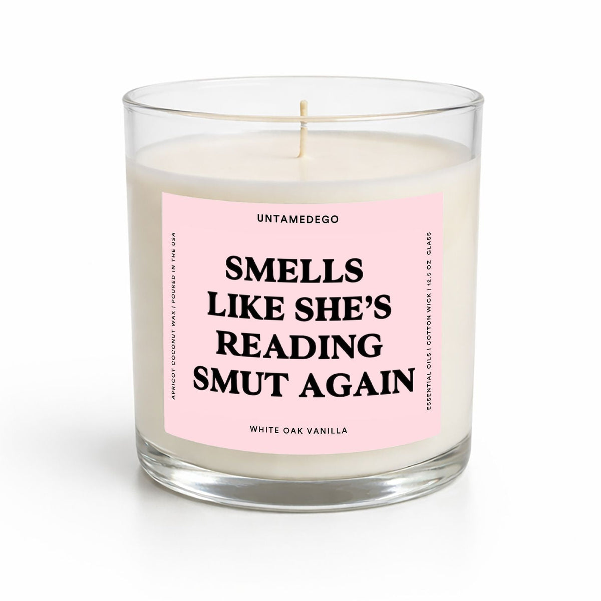 A white wax candle in a clear glass jar with a light pink label that reads 'SMELLS LIKE SHE'S READING SMUT AGAIN' in bold black text. The brand name 'UNTAMEDEGO' is visible at the top of the label.