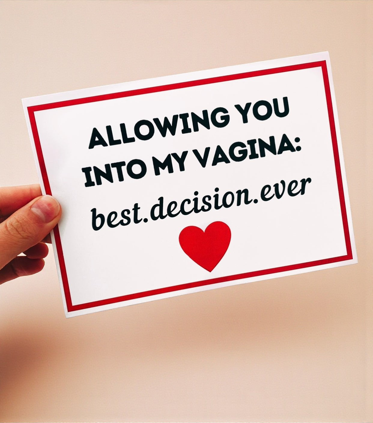 A hand holding a white greeting card with a red border and black text that reads “Allowing you into my vagina: best decision ever,” with a red heart printed below the text. The card is photographed against a warm cream-colored background.
