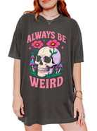 Graphic t-shirt with skull, flowers, and mushrooms design and text saying always be weird
