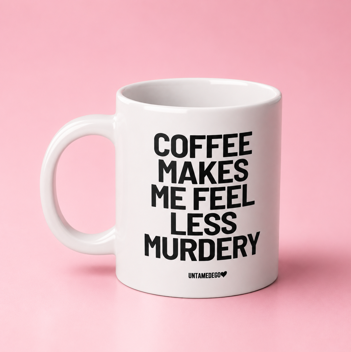 A white ceramic coffee mug with bold black text reading 'COFFEE MAKES ME FEEL LESS MURDERY' and a small 'UNTAMEDSO' logo at the bottom, set against a solid pink background.