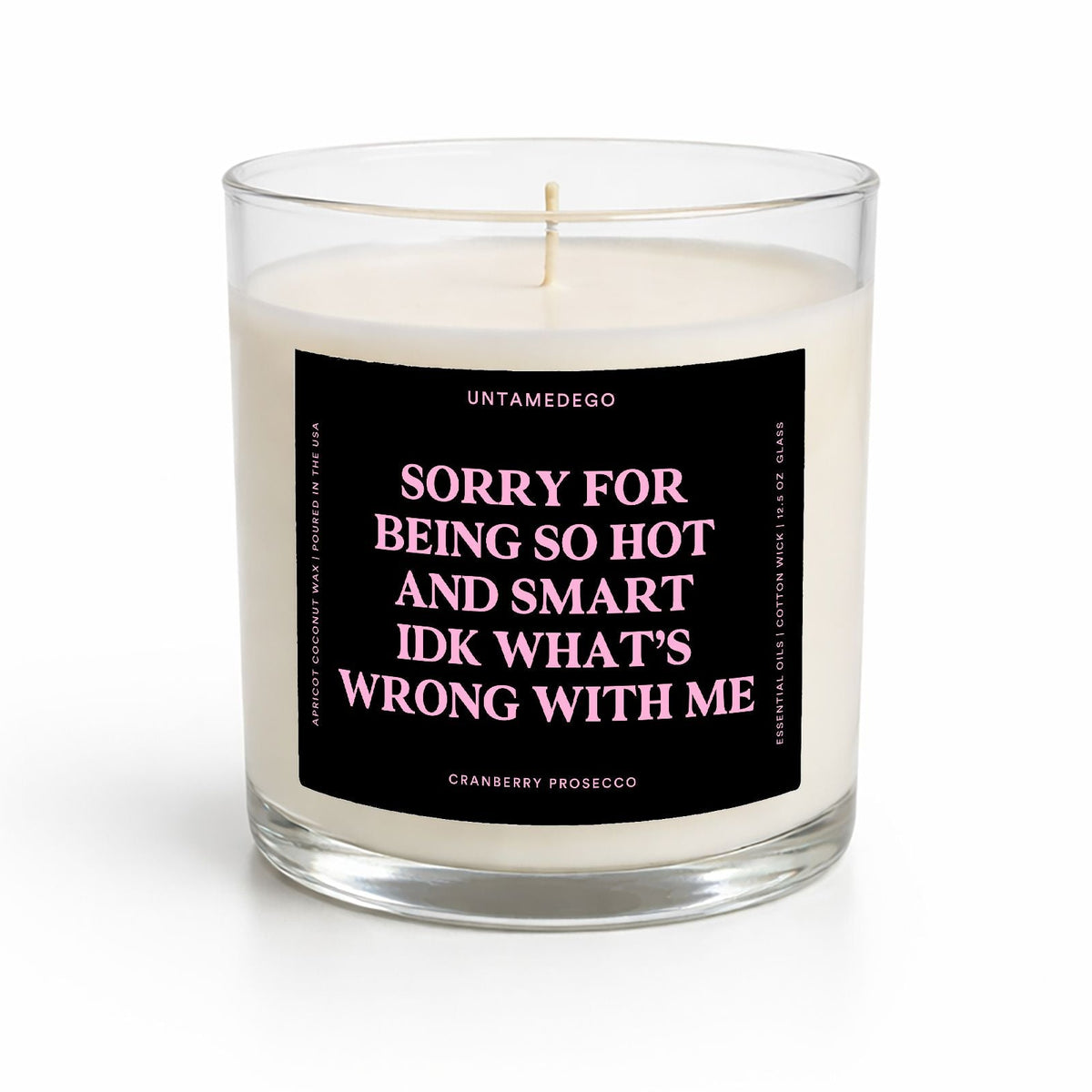 A single-wick candle in a clear glass jar with cream-colored wax and a black label featuring pink text that reads, 'SORRY FOR BEING SO HOT AND SMART IDK WHAT'S WRONG WITH ME.' The brand name 'UNTAMEDEGO' is visible at the top of the label.