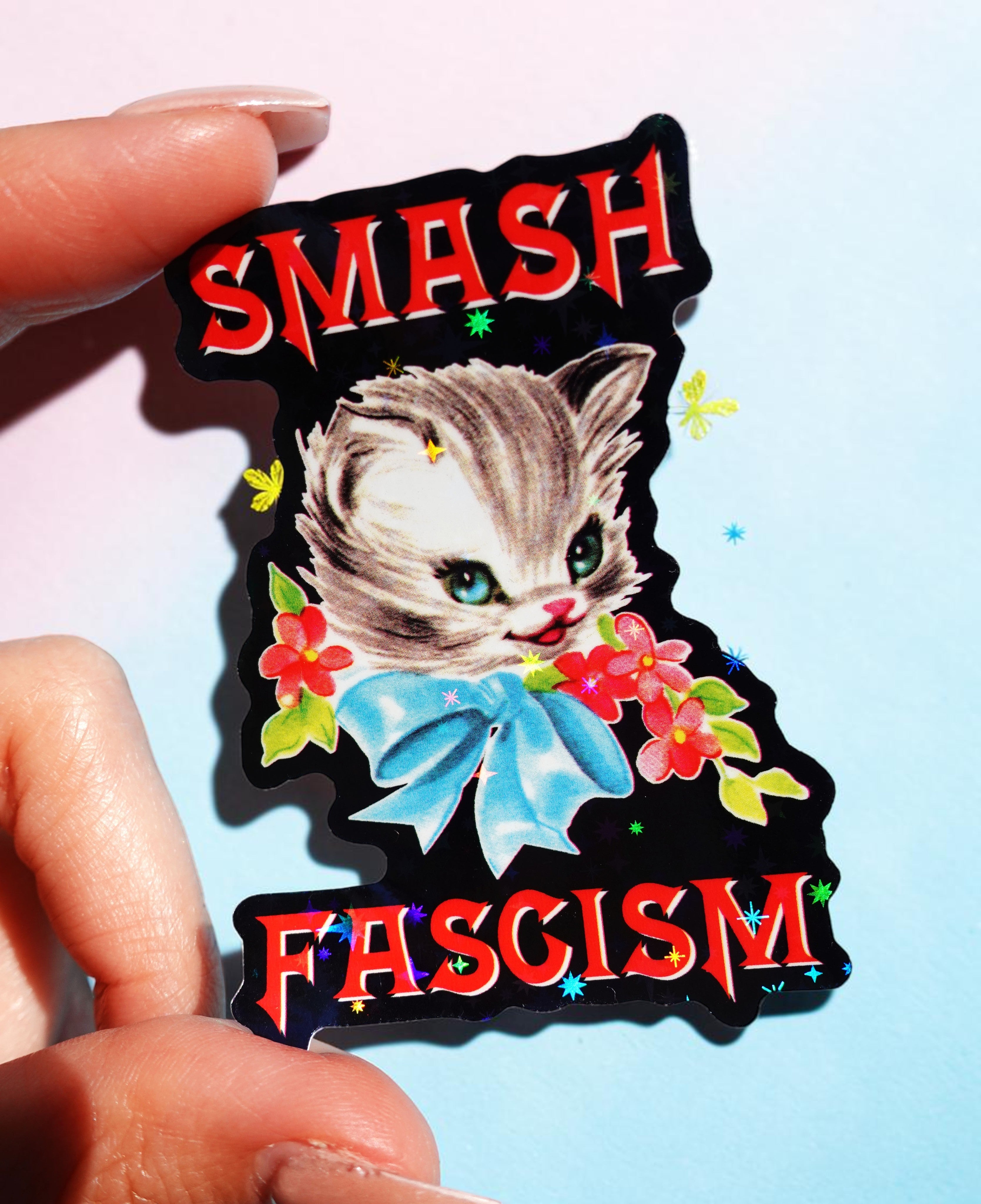Sticker with a cat design and 'SMASH FASCISM' text held by a hand against a light blue background.