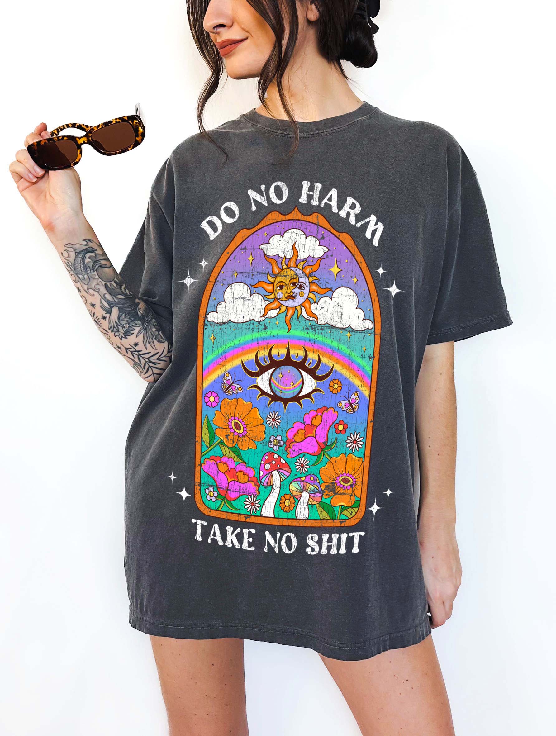 Person wearing a dark gray graphic t-shirt featuring a colorful illustrated scene with sun, rainbow, flowers, mushrooms, and the text Do No Harm Take No Shit, photographed against a light background