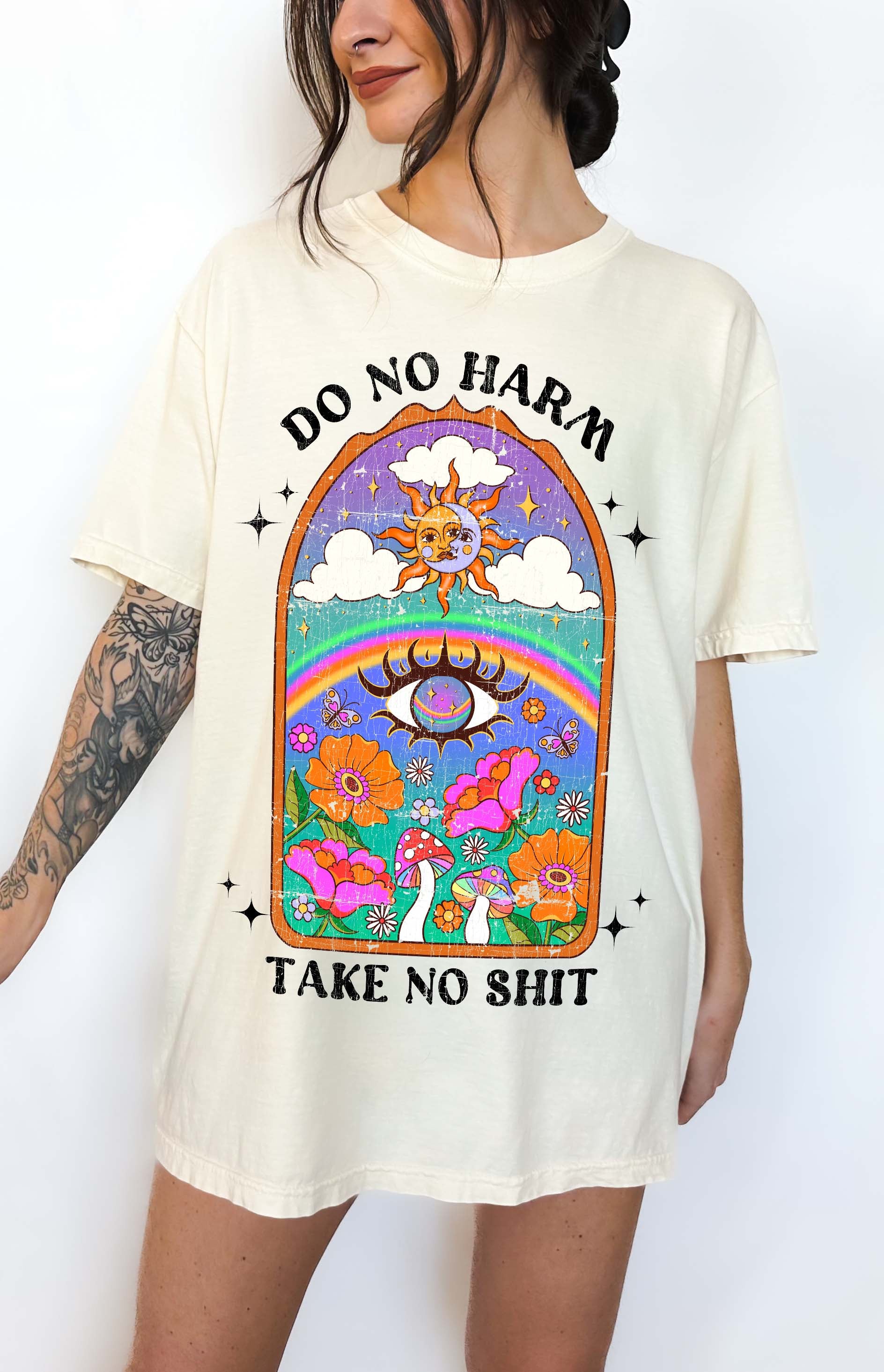 Person wearing Ivory graphic t-shirt featuring a colorful illustrated scene with sun, rainbow, flowers, mushrooms, and the text Do No Harm Take No Shit, photographed against a light background