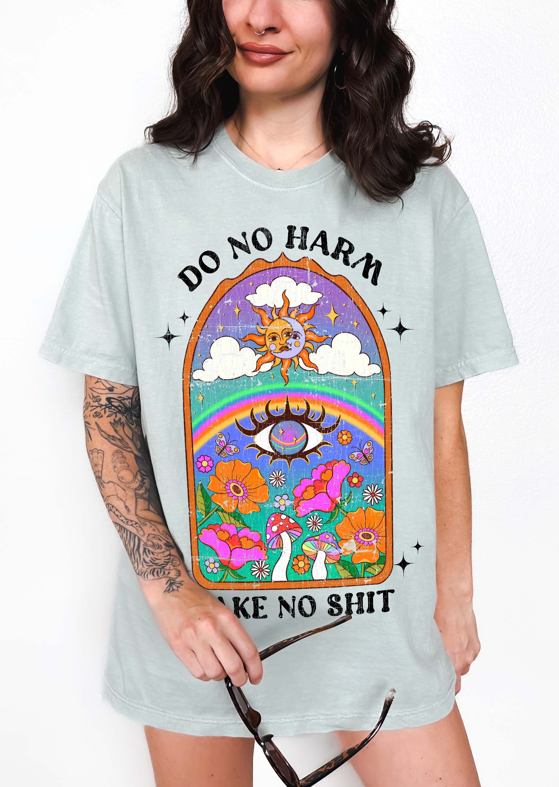 Person wearing a light blue graphic t-shirt featuring a colorful illustrated scene with sun, rainbow, flowers, mushrooms, and the text Do No Harm Take No Shit, photographed against a light background
