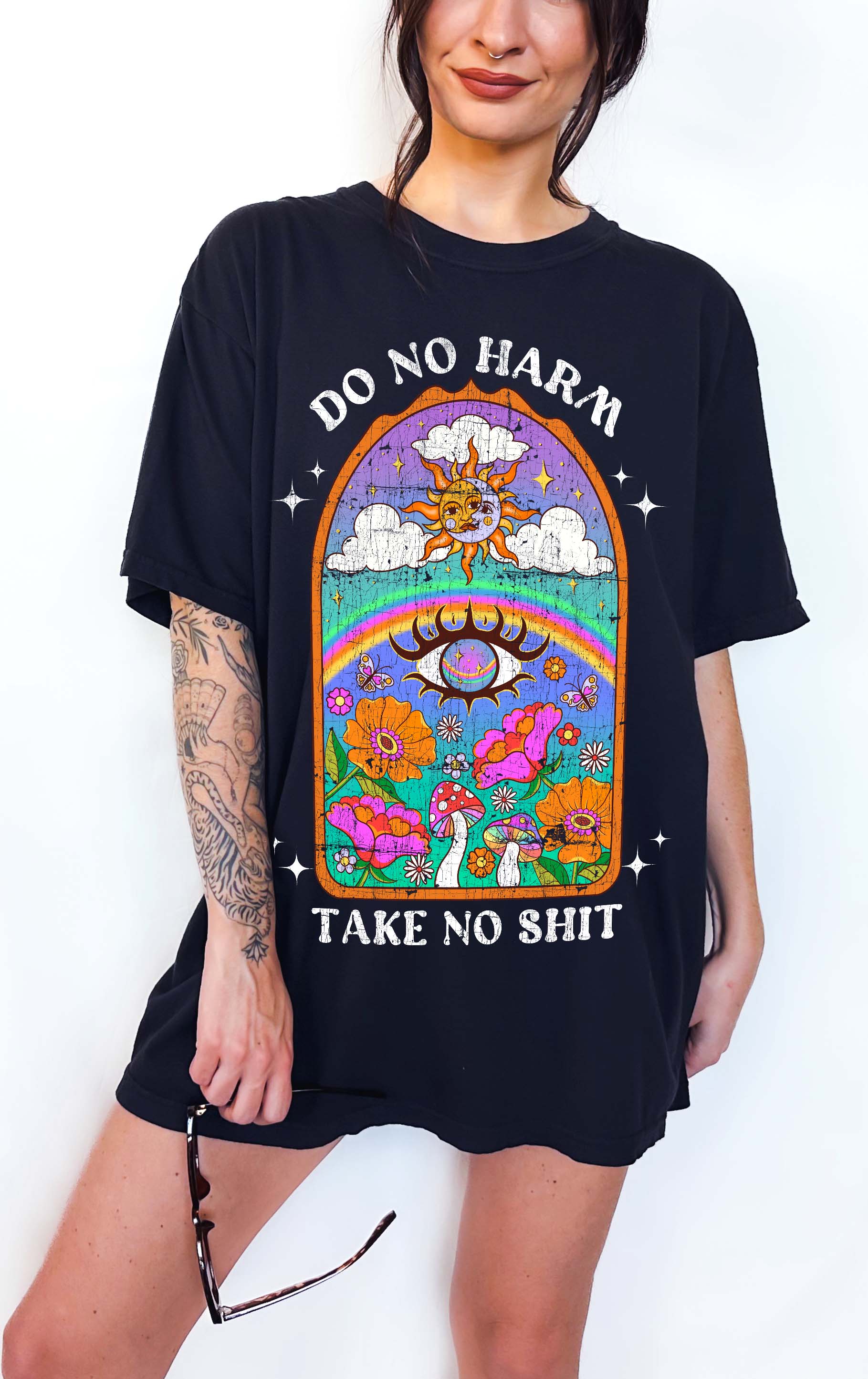 Person wearing a black graphic t-shirt featuring a colorful illustrated scene with sun, rainbow, flowers, mushrooms, and the text Do No Harm Take No Shit, photographed against a light background