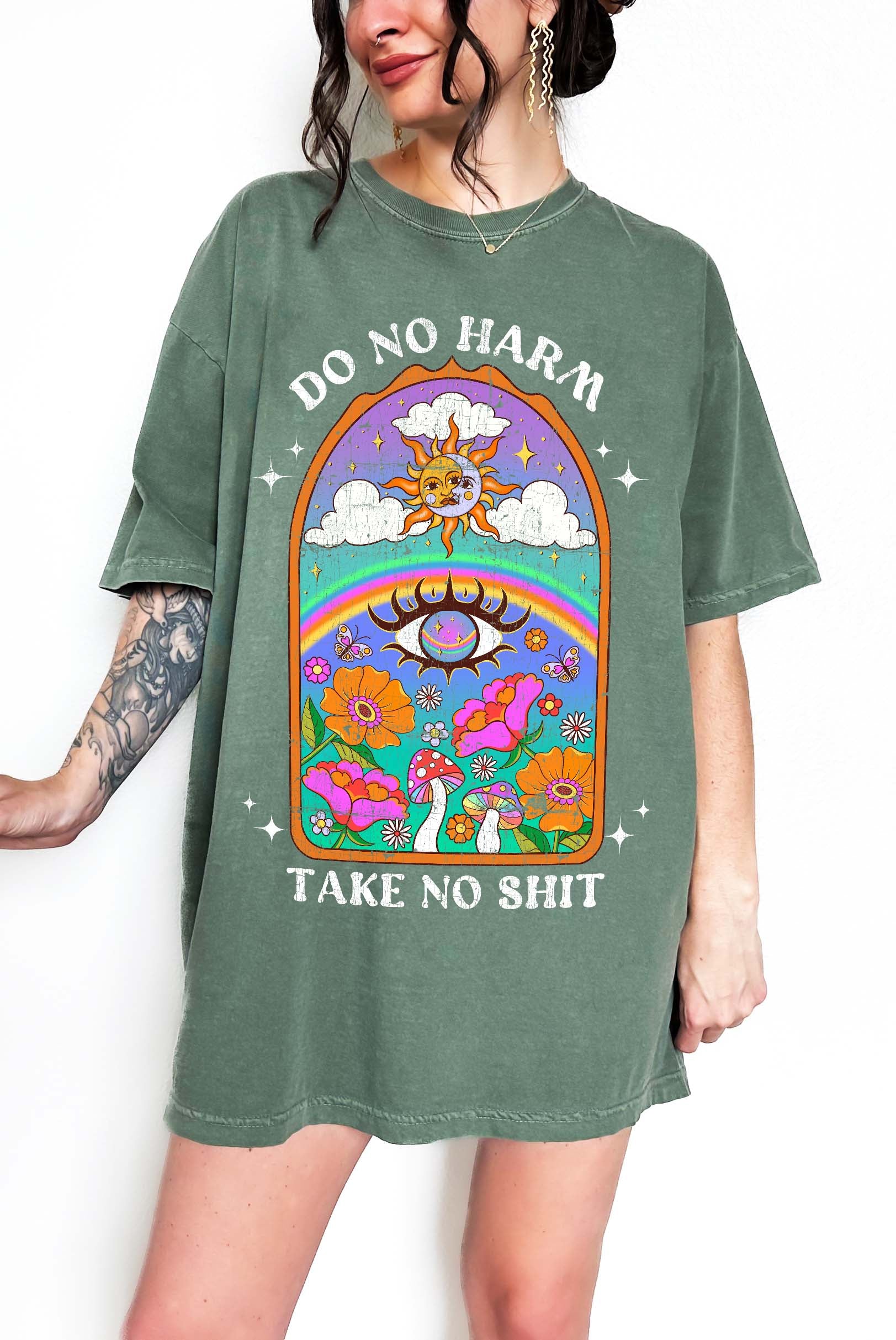 Person wearing a green graphic t-shirt featuring a colorful illustrated scene with sun, rainbow, flowers, mushrooms, and the text Do No Harm Take No Shit, photographed against a light background