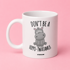 A white ceramic mug with a cartoon hippo graphic and the text 'DON'T BE A HIPPO-TWATAMUS' printed on it, set against a solid pink background.