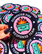 Round holographic glitter sticker reading "Dumpster Fire Response Team" held by a hand with dark glitter nails against a yellow background, surrounded by multiples of the same sticker on a dark surface.