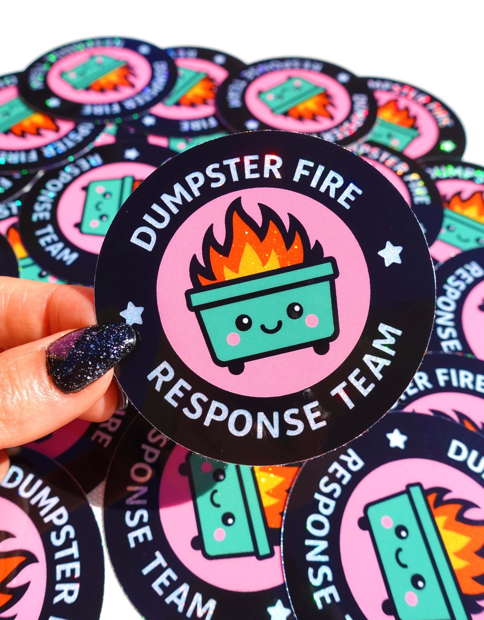 Round holographic glitter sticker reading "Dumpster Fire Response Team" held by a hand with dark glitter nails against a yellow background, surrounded by multiples of the same sticker on a dark surface.