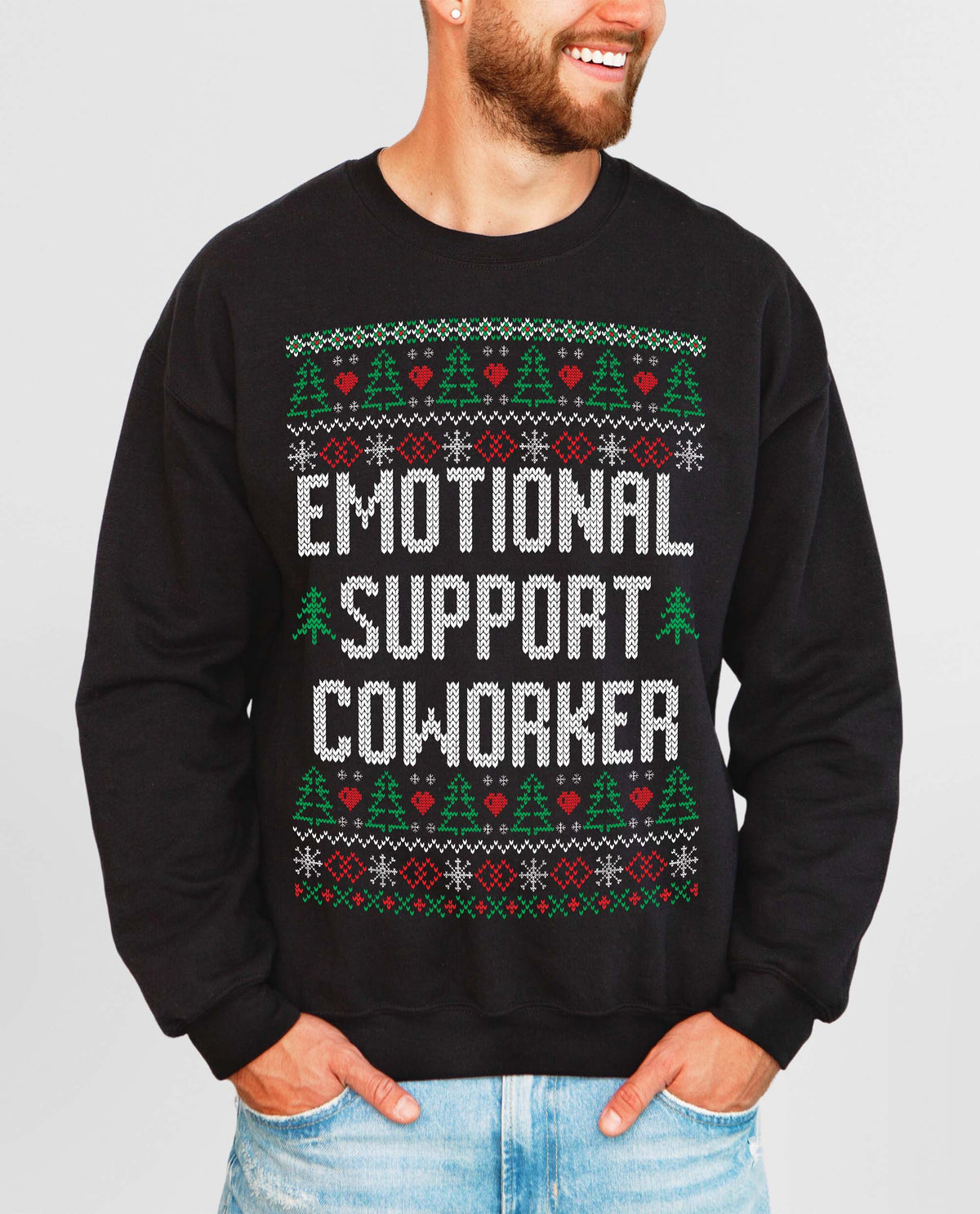 A man wearing a black crewneck sweatshirt with a festive Christmas-themed graphic that reads 'EMOTIONAL SUPPORT COWORKER' in white pixelated text, flanked by green and red holiday patterns, paired with light blue jeans against a solid gray background.