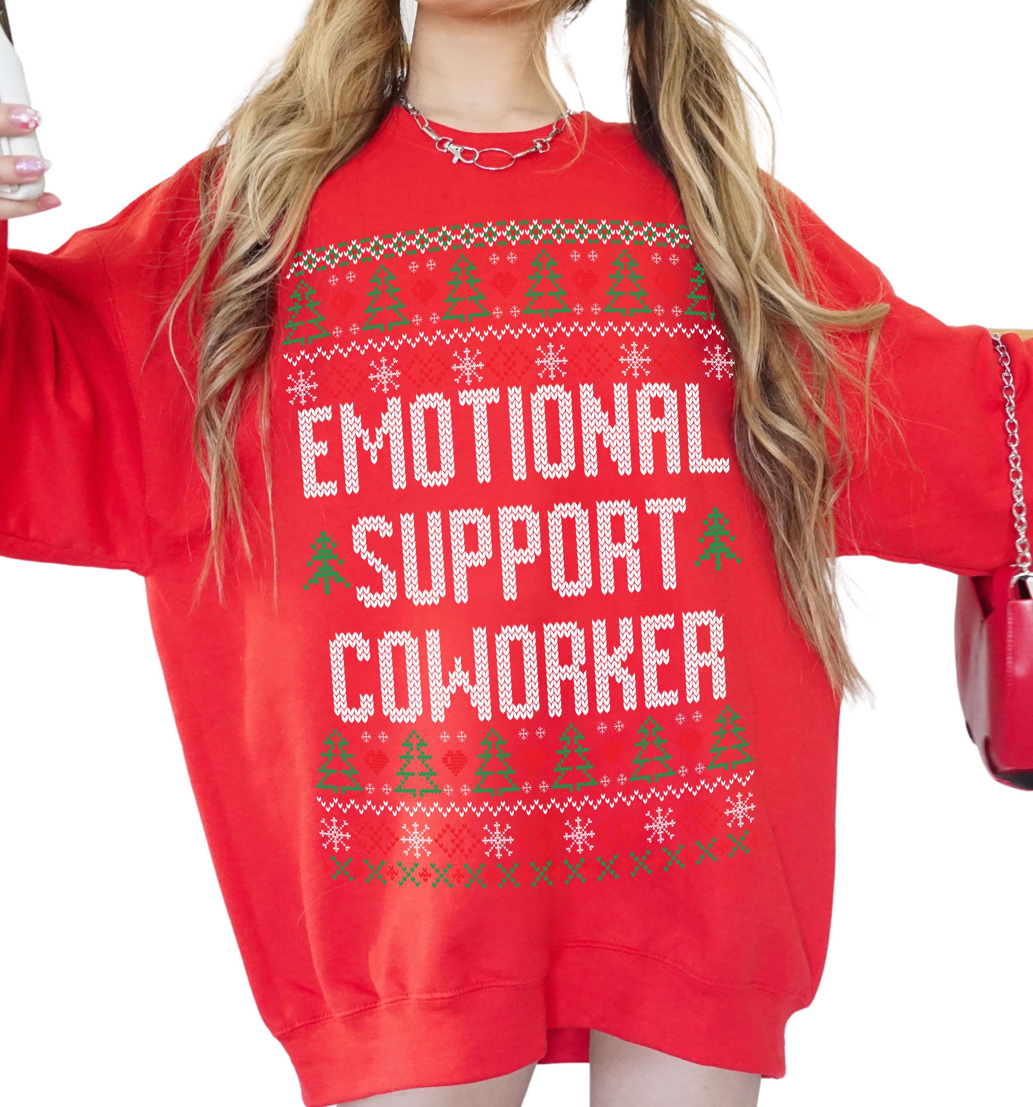 A woman wearing a bright red crewneck sweatshirt with a white and green pixelated holiday sweater design that reads 'EMOTIONAL SUPPORT COWORKER'.