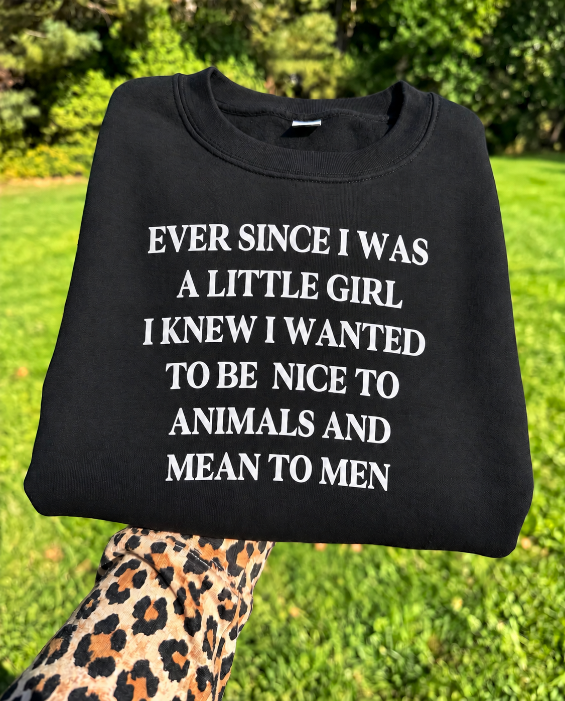 A black crewneck sweatshirt with bold white text reading 'EVER SINCE I WAS A LITTLE GIRL I KNEW I WANTED TO BE NICE TO ANIMALS AND MEAN TO MEN', held up by a leopard-print sleeve against a blurred green grass and trees background.