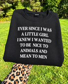 A black crewneck sweatshirt with bold white text reading 'EVER SINCE I WAS A LITTLE GIRL I KNEW I WANTED TO BE NICE TO ANIMALS AND MEAN TO MEN', held up by a leopard-print sleeve against a blurred green grass and trees background.