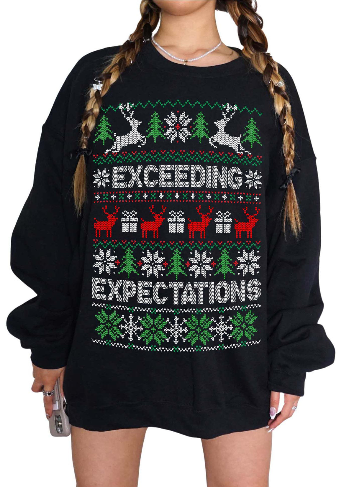 A person wearing an oversized black long-sleeve sweatshirt with a festive Christmas sweater print featuring reindeer, trees, snowflakes, and the words 'EXCEEDING EXPECTATIONS' in white, green, and red.