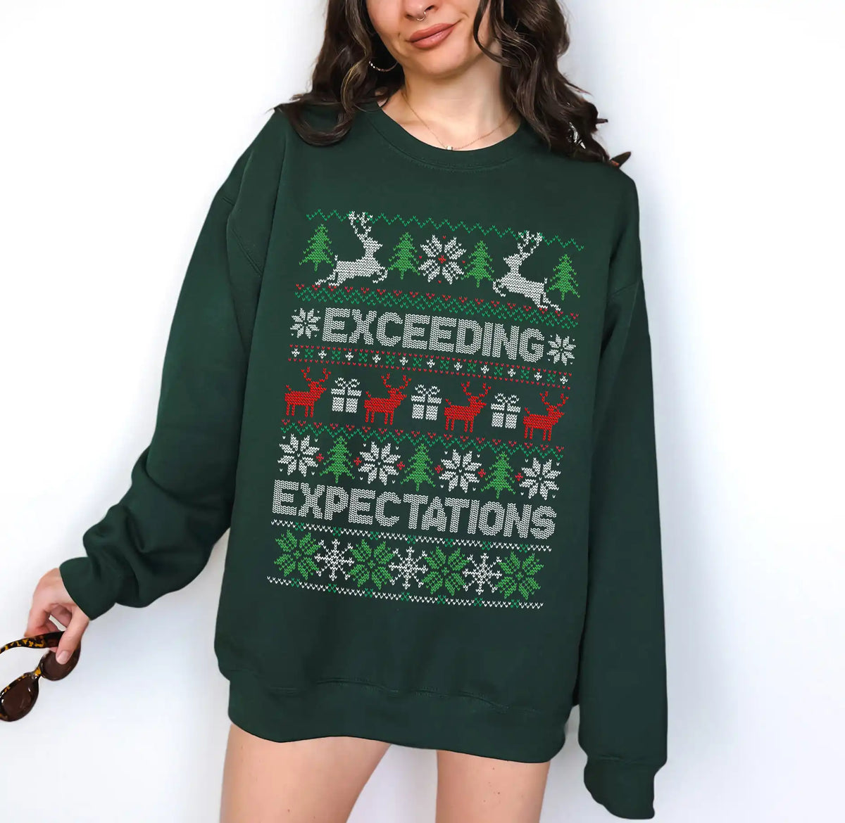 A woman wearing a dark green crewneck sweatshirt with a festive Christmas pattern featuring reindeer, snowflakes, and the text 'EXCEEDING EXPECTATIONS' in white and red.