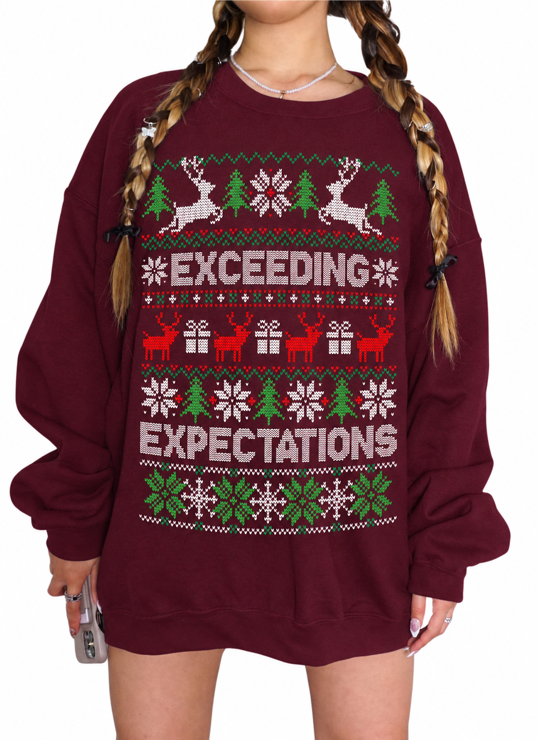 A person wearing an oversized crewneck sweatshirt in burgundy with a festive Christmas knit pattern featuring reindeer, snowflakes, trees, and the words 'EXCEEDING EXPECTATIONS' in white, red, and green.
