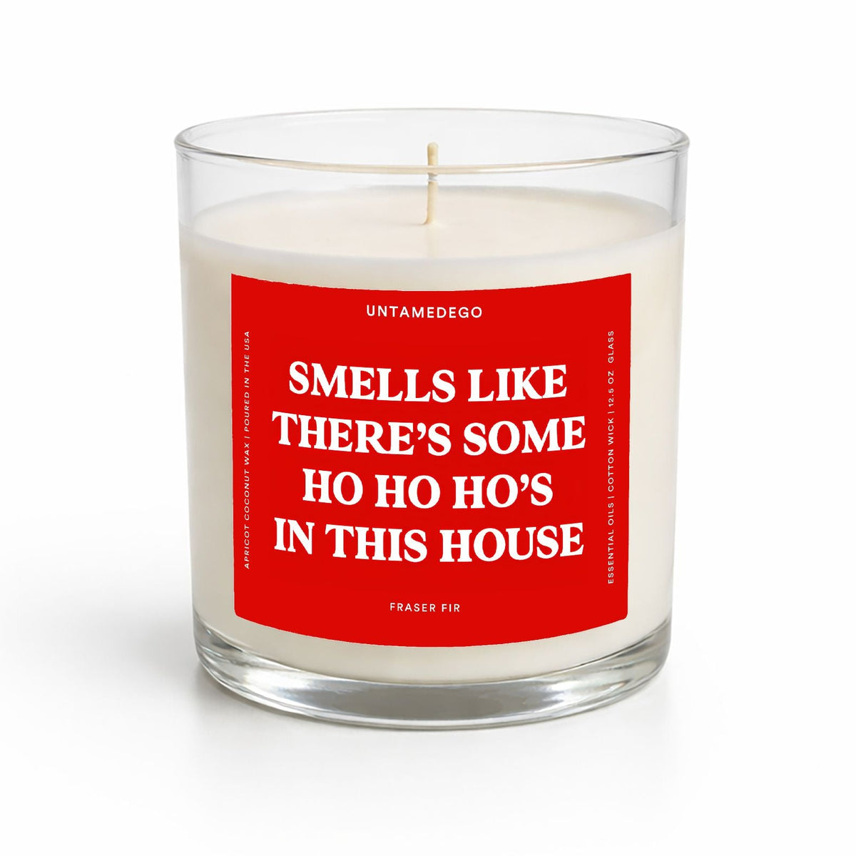 A clear glass jar candle with white wax and a prominent red label featuring white text that reads, 'SMELLS LIKE THERE'S SOME HO HO HO'S IN THIS HOUSE.' The brand name 'UNTAMEDEGO' is visible at the top of the label.