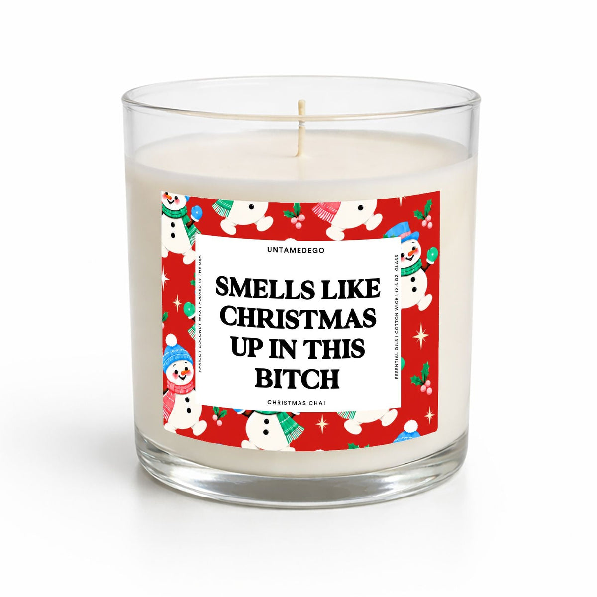 A clear glass jar candle with white wax and a humorous Christmas-themed label that reads 'SMELLS LIKE CHRISTMAS UP IN THIS BITCH.' The label features a red border with cartoon snowman illustrations and the brand name Untamed Co.