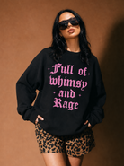A woman modeling a black crewneck sweatshirt with pink gothic text reading 'Full of whimsy and Rage', paired with leopard-print shorts, against a warm brown studio backdrop.