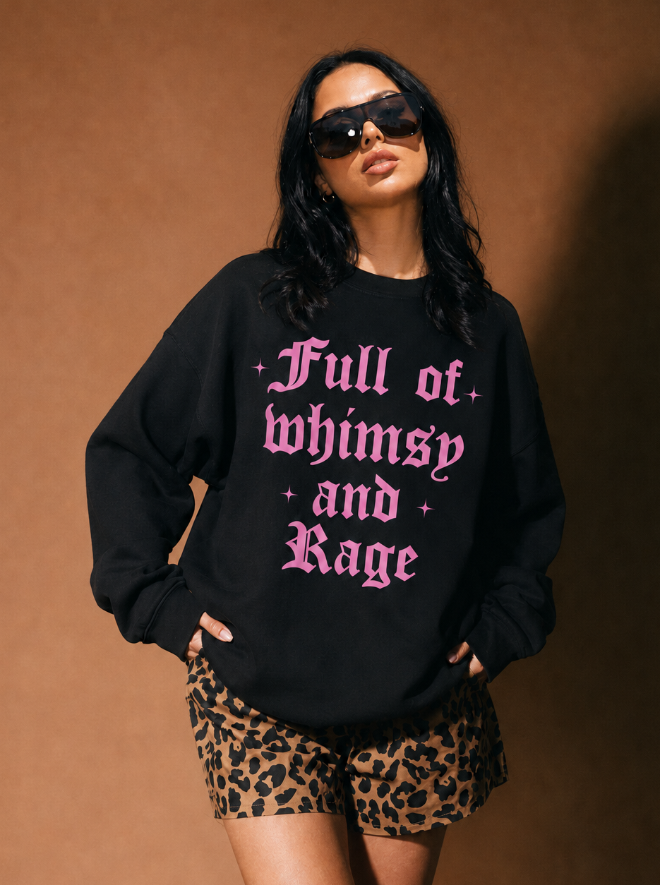 A woman modeling a black crewneck sweatshirt with pink gothic text reading 'Full of whimsy and Rage', paired with leopard-print shorts, against a warm brown studio backdrop.