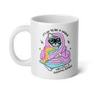 A white ceramic mug featuring a cartoon illustration of a cat wrapped in a pink blanket with a rainbow design, wearing black sunglasses, with the text 'IT'S OK TO BE A SADDIE YOU STILL A FUCKING SADDIE' printed on the side.