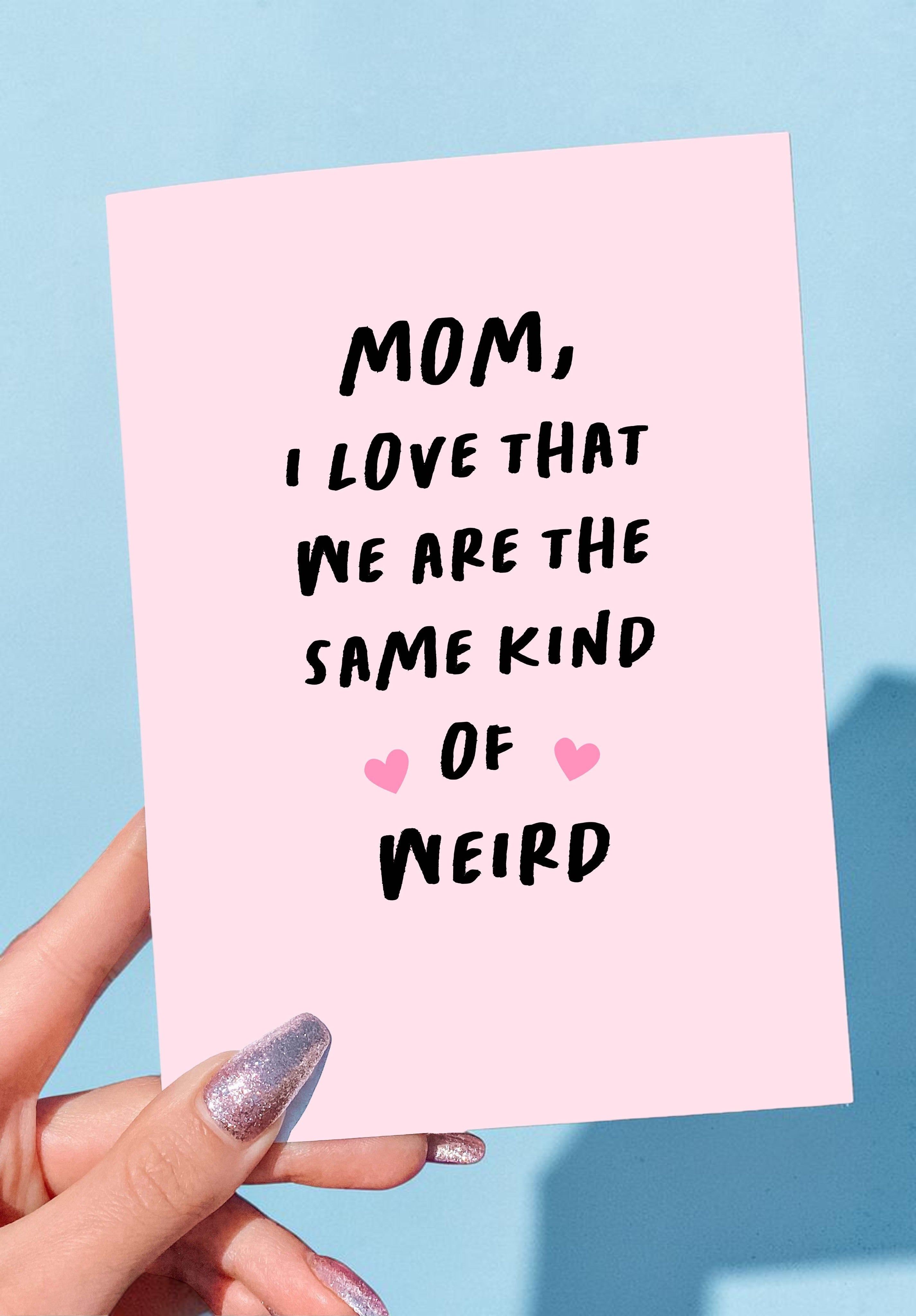 Pink card with text 'Mom, I love that we are the same kind of weird' held by a hand against a blue background