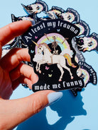 Holographic glitter vinyl sticker showing a skeleton in a party hat riding a unicorn under a rainbow with butterflies and clouds. Bold white text reads At Least My Trauma Made Me Funny.