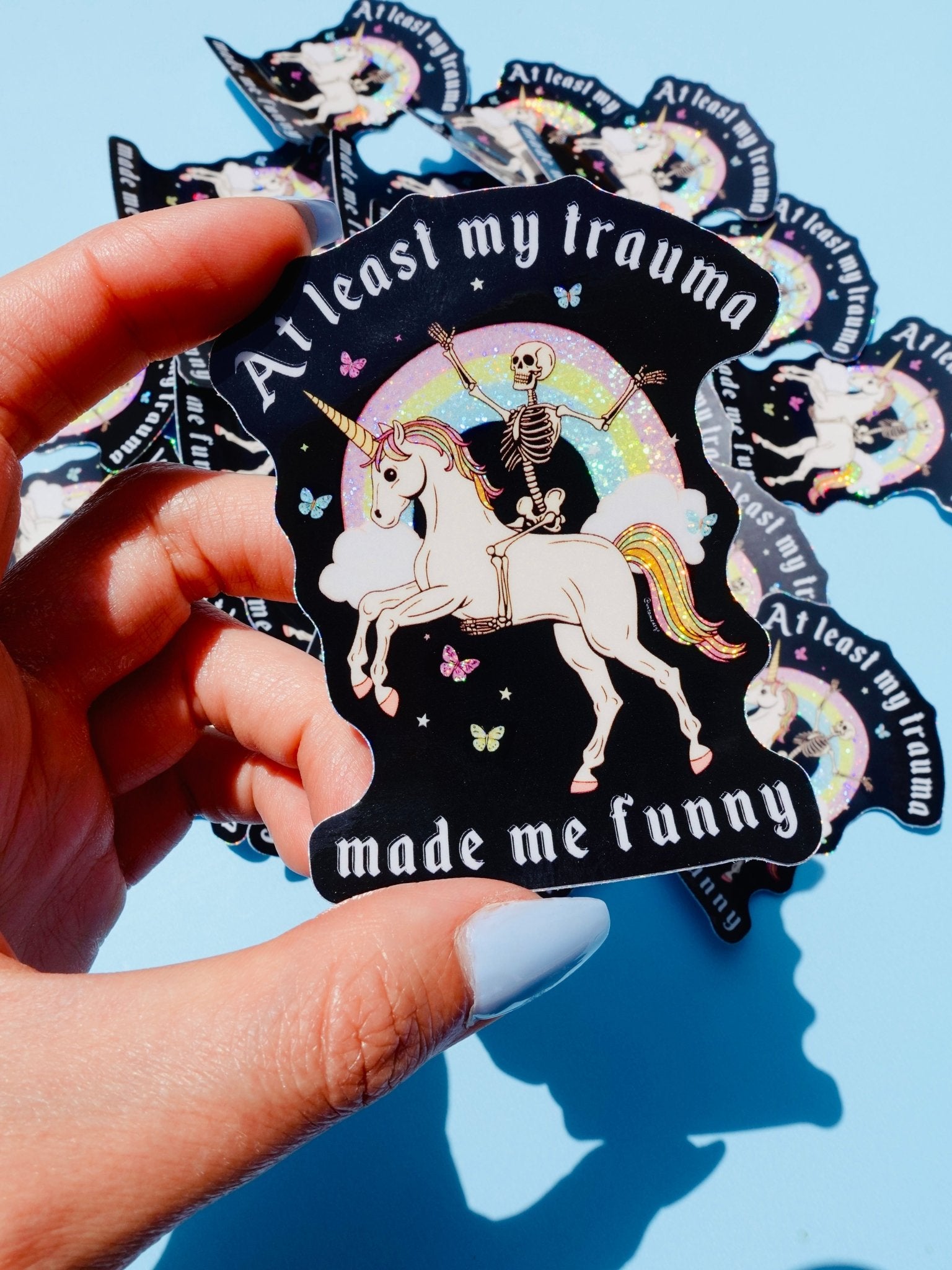 Holographic glitter vinyl sticker showing a skeleton in a party hat riding a unicorn under a rainbow with butterflies and clouds. Bold white text reads At Least My Trauma Made Me Funny.