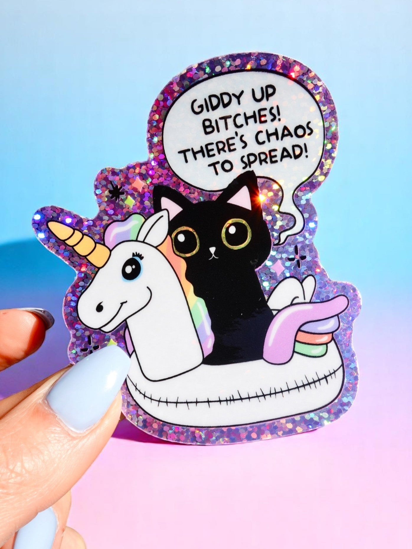 Die-cut holographic vinyl sticker featuring a black cat riding a purple unicorn pool float. Speech bubble text reads: Giddy Up Bitches! There's chaos to spread. Purple and pink background with glitter effect