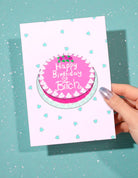 Birthday greeting card featuring a pink illustrated cake with the words Happy Birthday on the cake and small heart graphics on a pastel background, photographed against a teal glitter background