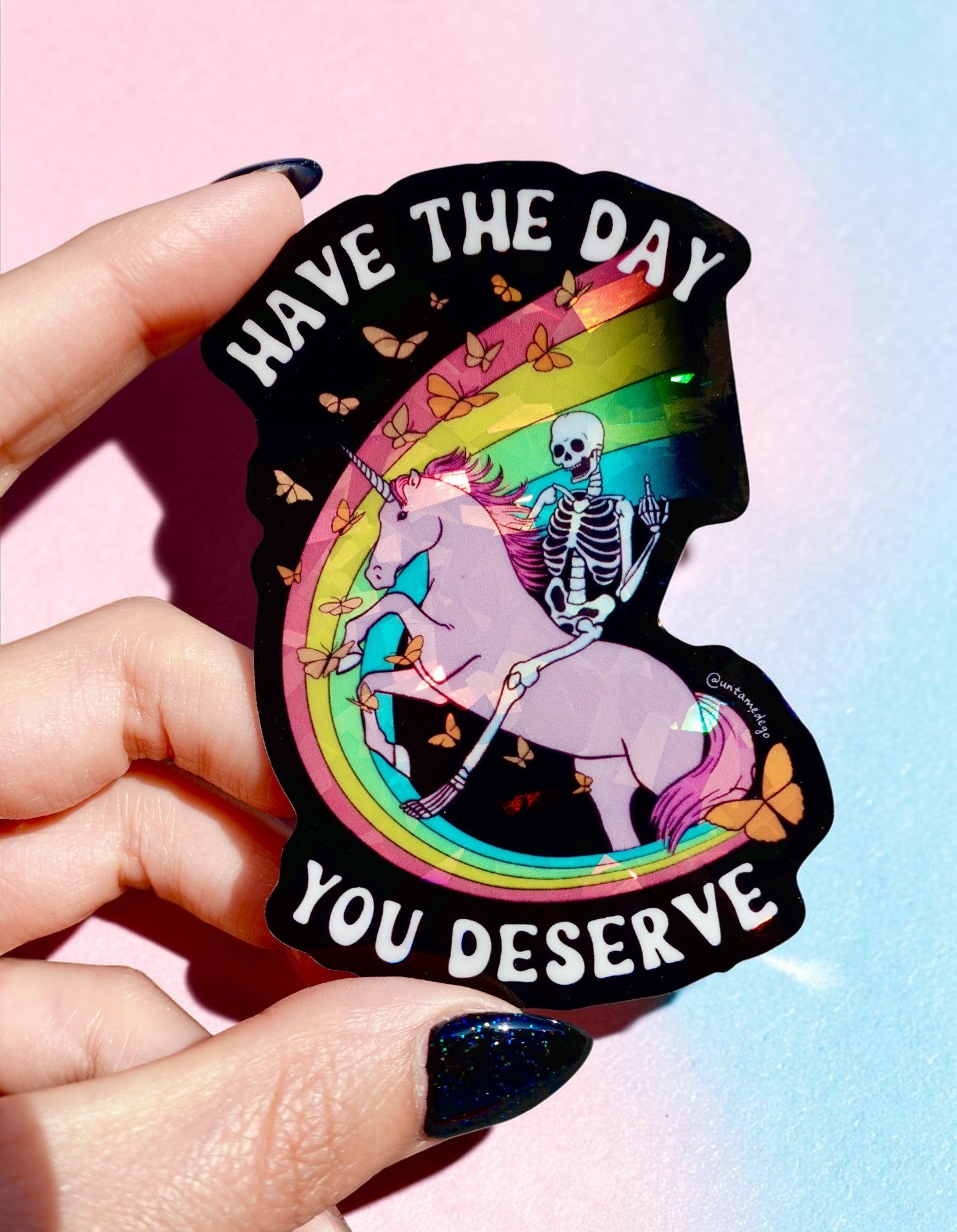 Funny holographic sticker with skeleton riding a unicorn and text “have the day you deserve” cute dark humor vinyl sticker