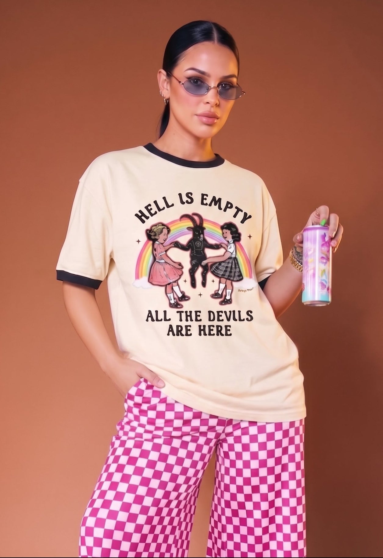 A woman modeling a cream graphic t-shirt with black trim, pink and white checkered wide-leg pants, and lime green sandals, holding a pink spray can against a solid brown studio background.