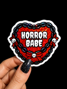 A hand with glittery black nail polish holds a die-cut sticker featuring a red glitter heart, black chain border, white skulls, and the text 'HORROR BABE' in white. The background is solid black.