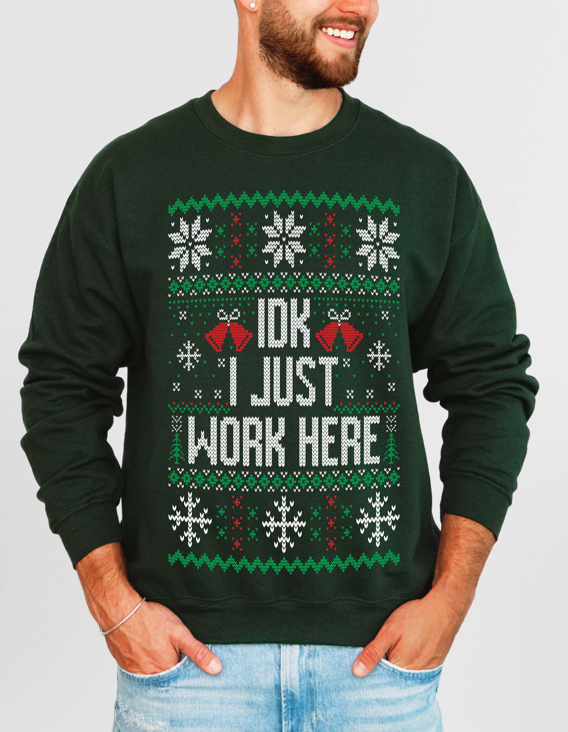 A man wearing a dark green crewneck sweatshirt with a festive Christmas-themed graphic that reads 'IDK I JUST WORK HERE' in white text, flanked by red jingle bells and decorated with snowflakes and green/red patterns.