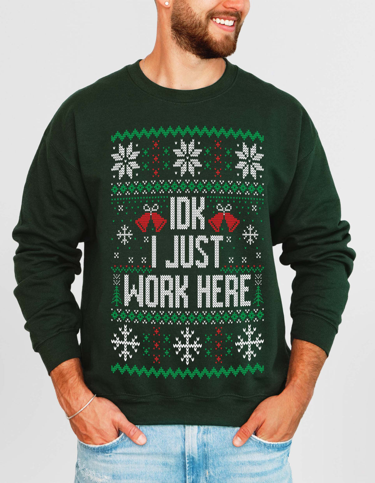 A man wearing a dark green crewneck sweatshirt with a festive Christmas-themed graphic that reads 'IDK I JUST WORK HERE' in white text, flanked by red jingle bells and decorated with snowflakes and green/red patterns.