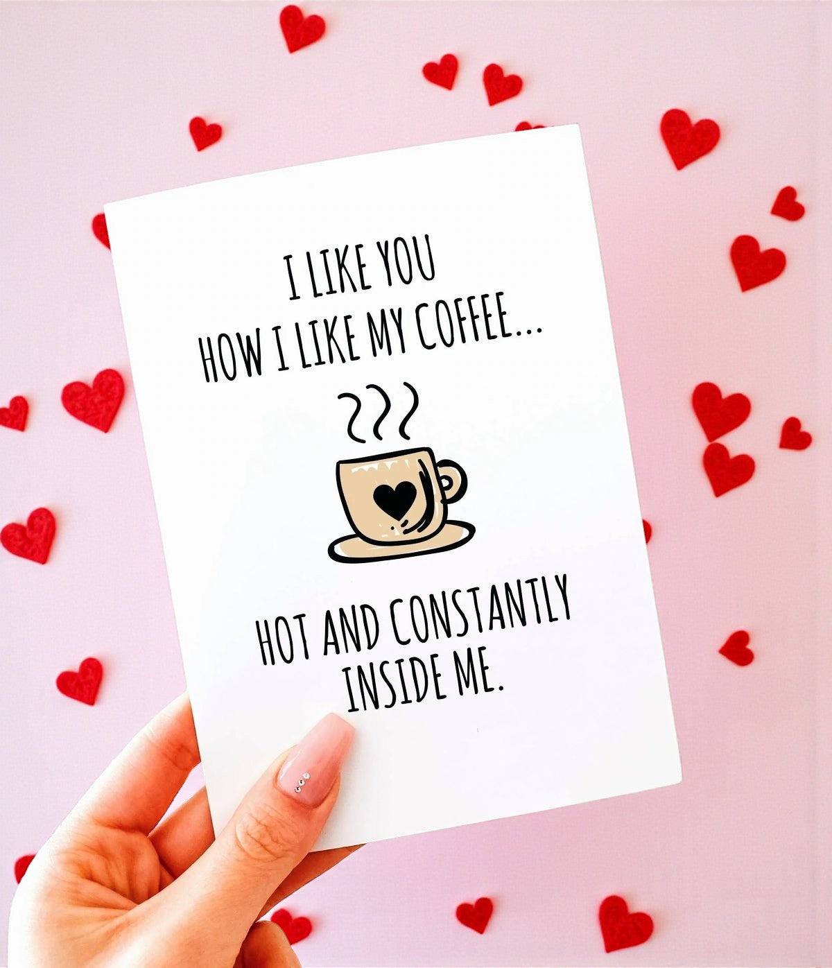 A hand holding a white greeting card with black text that reads “I like you how I like my coffee… hot and constantly inside me,” with a small illustrated coffee cup featuring a heart. The background is soft blush pink with scattered red heart shapes.