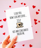 A hand holding a white greeting card with black text that reads “I like you how I like my coffee… hot and constantly inside me,” with a small illustrated coffee cup featuring a heart. The background is soft blush pink with scattered red heart shapes.