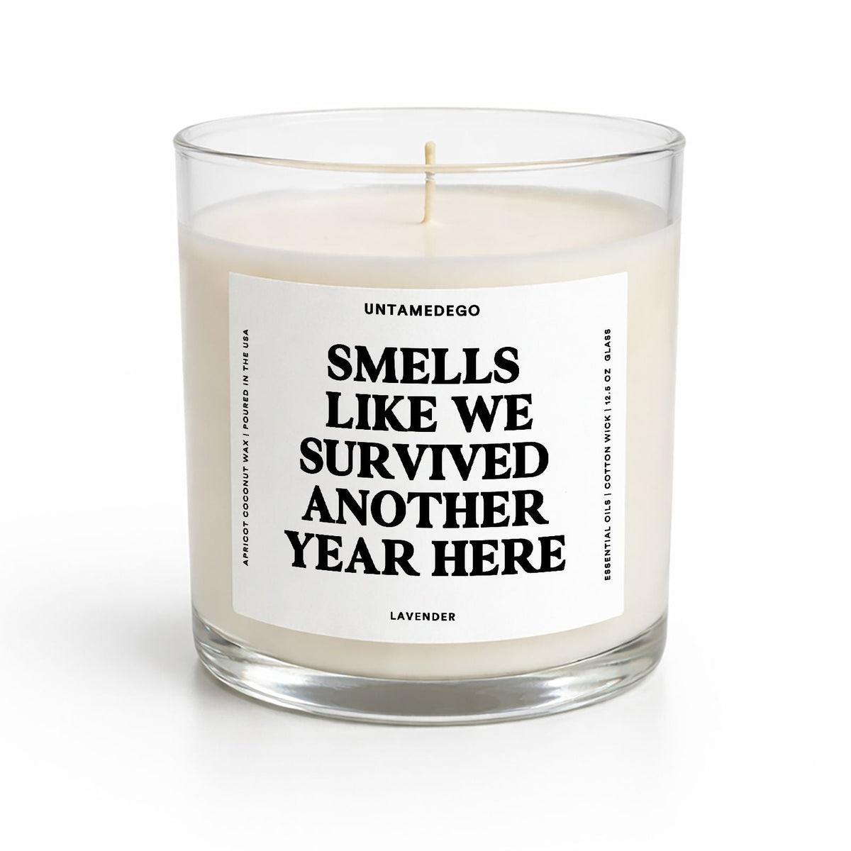 A clear glass jar candle with cream-colored wax and a single wick, featuring a white label with bold black text that reads 'SMELLS LIKE WE SURVIVED ANOTHER YEAR HERE' and 'UNTAMEDOGO' at the top, on a clean white background.