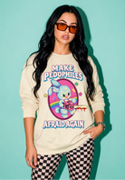 A woman modeling a cream-colored crewneck sweatshirt with a colorful graphic of a cartoon bunny and the text 'MAKE PEDOPHILES AFRAID AGAIN'. She is wearing orange-tinted sunglasses, a watch, and black-and-white checkered pants against a solid teal background.