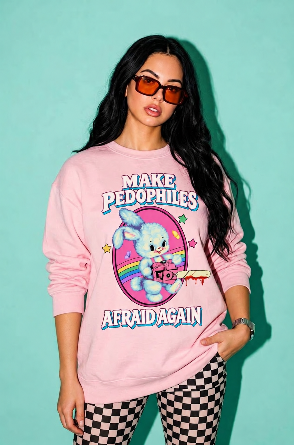 A woman modeling a light pink crewneck sweatshirt with a graphic print that reads 'MAKE PEDOPHILES AFRAID AGAIN' and features a cartoon bunny holding a knife. She is wearing orange-tinted sunglasses and black-and-white checkered pants against a solid mint green background.