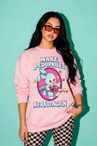A woman modeling a light pink crewneck sweatshirt with a graphic print that reads 'MAKE PEDOPHILES AFRAID AGAIN' and features a cartoon bunny holding a knife. She is wearing orange-tinted sunglasses and black-and-white checkered pants against a solid mint green background.