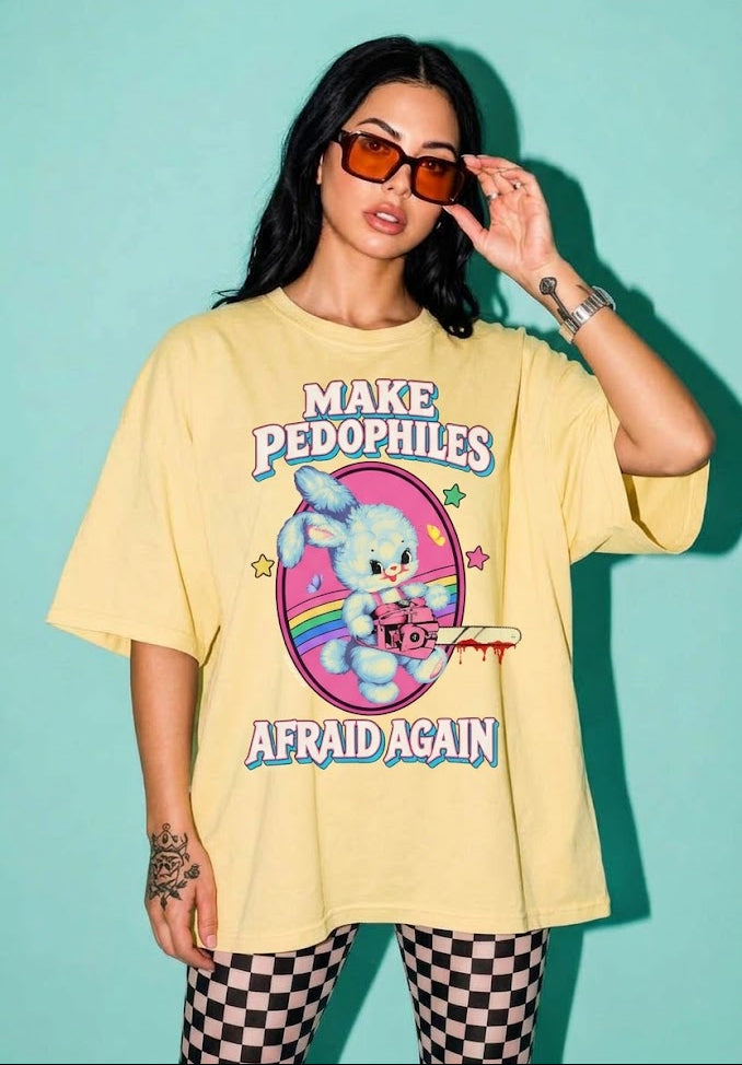 A woman modeling an oversized pale yellow graphic t-shirt with the text 'MAKE PEDOPHILES AFRAID AGAIN' and a cartoon bunny illustration, paired with black and white checkered pants against a solid teal background.