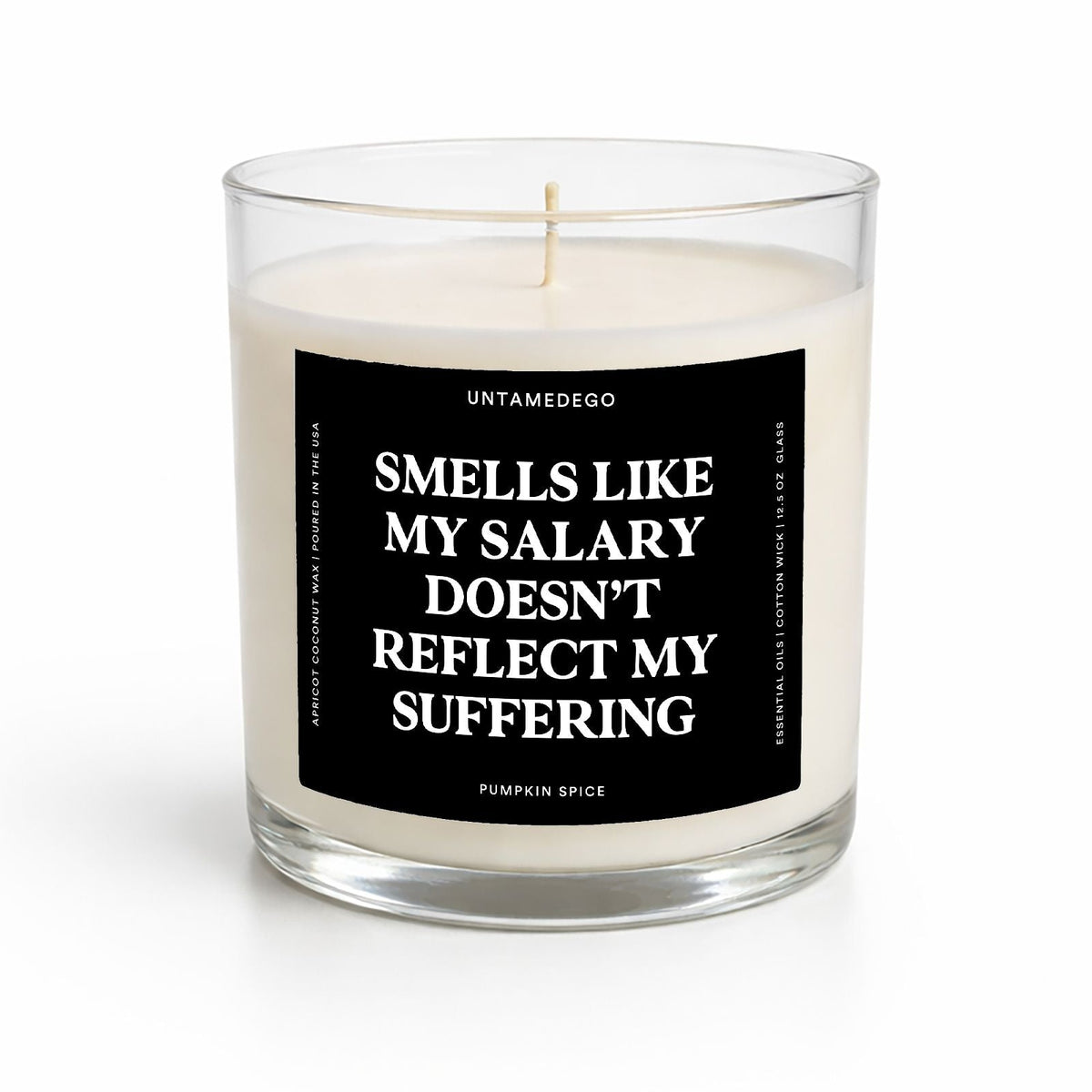 A clear glass jar candle with white wax and a black label featuring white text that reads, 'SMELLS LIKE MY SALARY DOESN'T REFLECT MY SUFFERING' with the brand name 'UNTAMEDEGO' at the top and 'PUMPKIN SPICE' at the bottom.