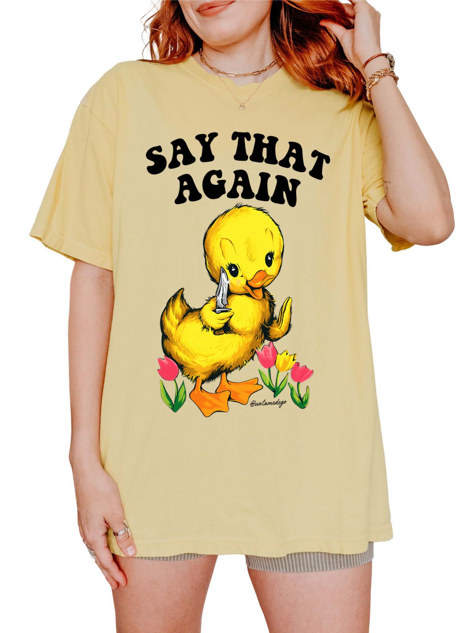 A person wearing a yellow crew neck t-shirt with a graphic that says 'SAY THAT AGAIN' and features an illustration of a duck with a flower. The t-shirt has a relaxed fit.