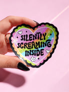 Holographic heart sticker reading "Silently Screaming Inside" held by a hand with dark nails against a pink background, showing pastel rainbow colors and black scalloped border detail.