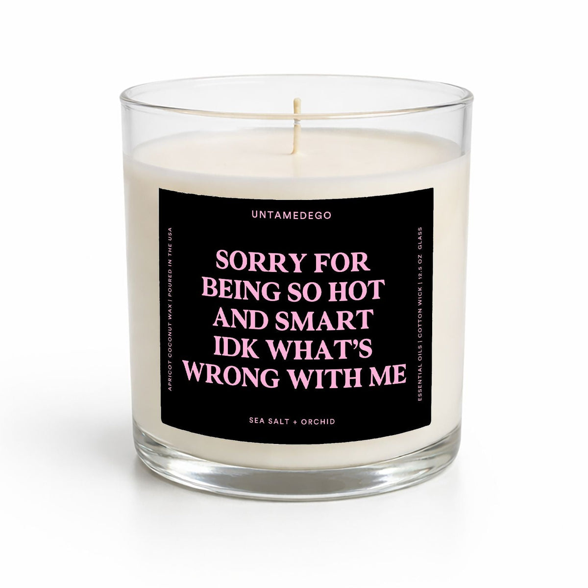 A sea salt + orchid scented candle in a clear glass jar with a black label featuring pink text that reads 'SORRY FOR BEING SO HOT AND SMART IDK WHAT'S WRONG WITH ME' by Untamedeo.
