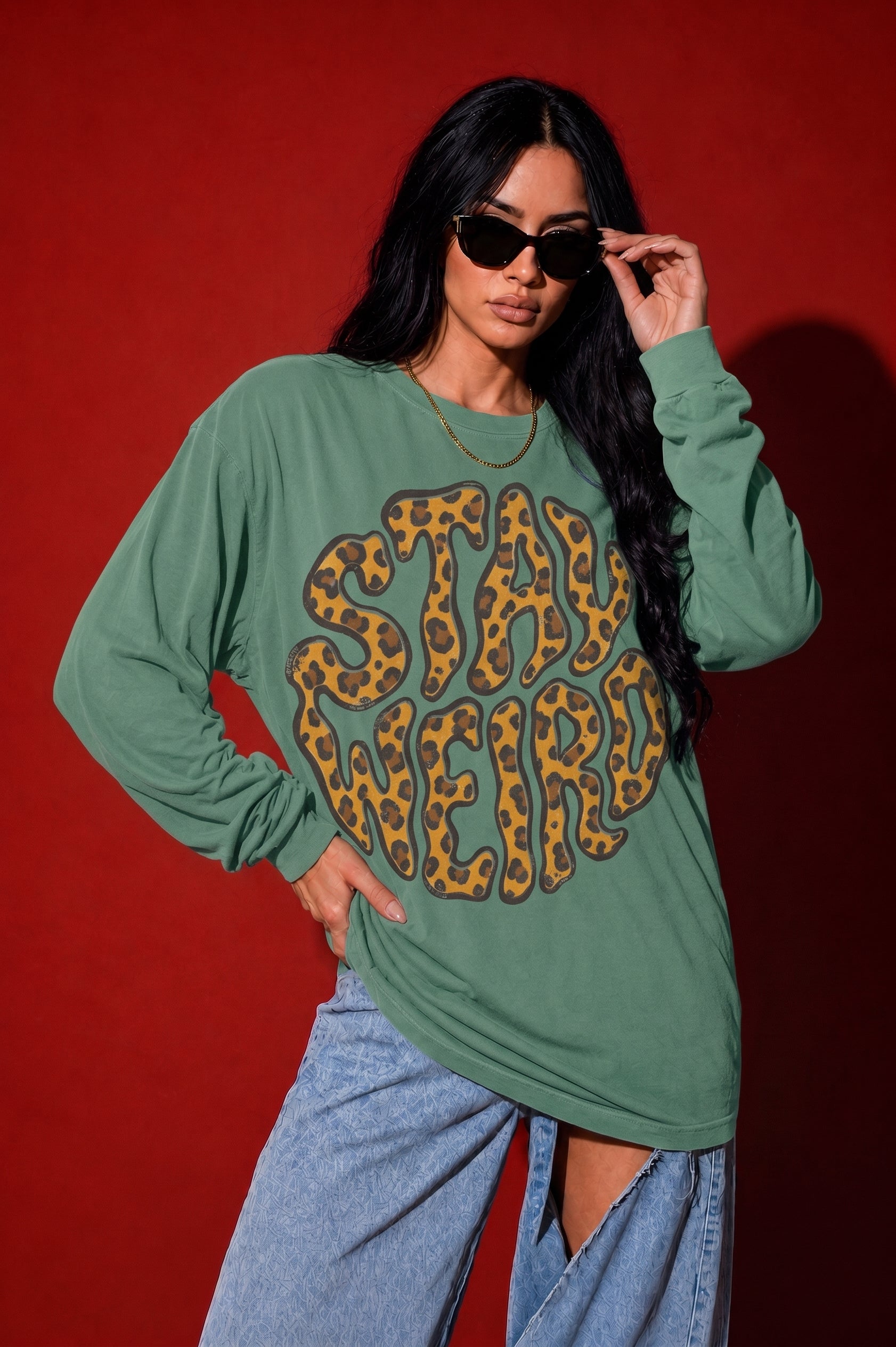 A woman modeling an oversized sage green long-sleeve t-shirt with a large leopard-print graphic that reads 'STAY WEIRD' across the front, paired with light-wash jeans against a solid red studio background.